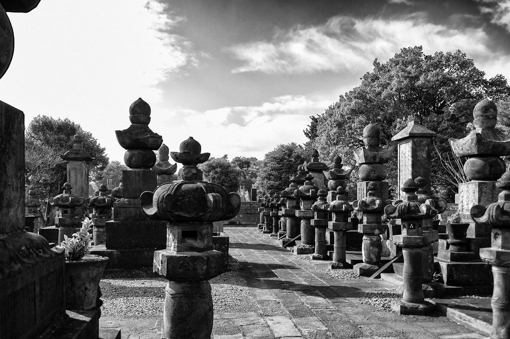 Monochrome photo of Yanaka cemetery in Yanaka, Tokyo, showing two rows of large monuments, some in the shape of people, separated by a paving stone pathway, against a cloud-and-sky background.