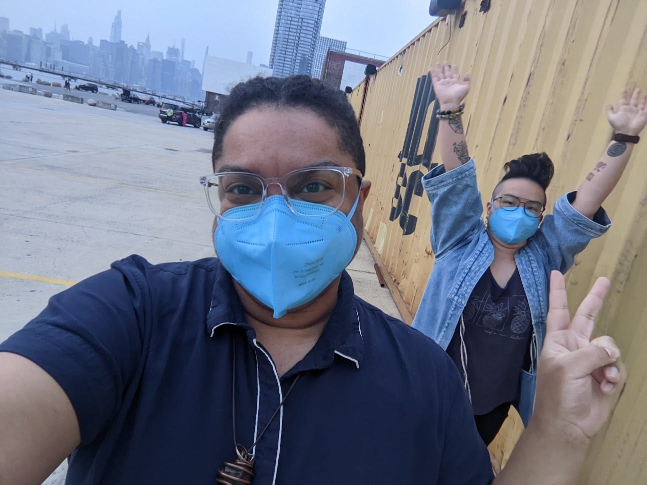 two queer humans wearing glasses and blue N95 masks outside. one is making a peace sign and one has their arms in the air.