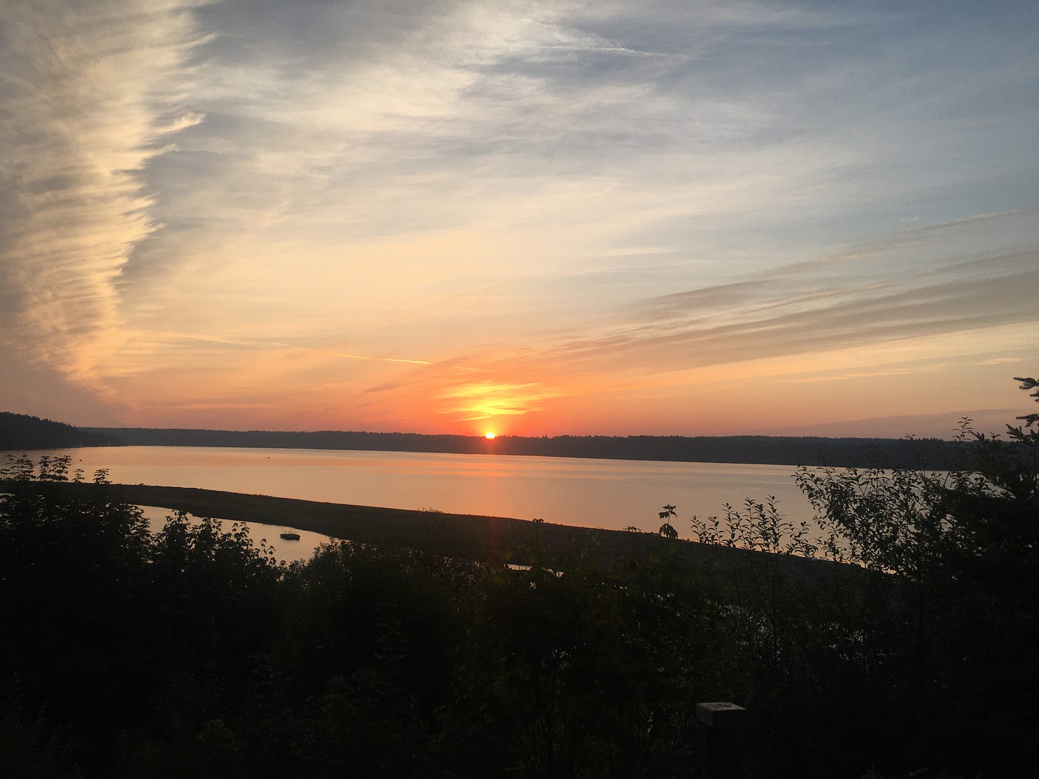 The sun is peeking over the distant tops of trees as seen across an inlet.