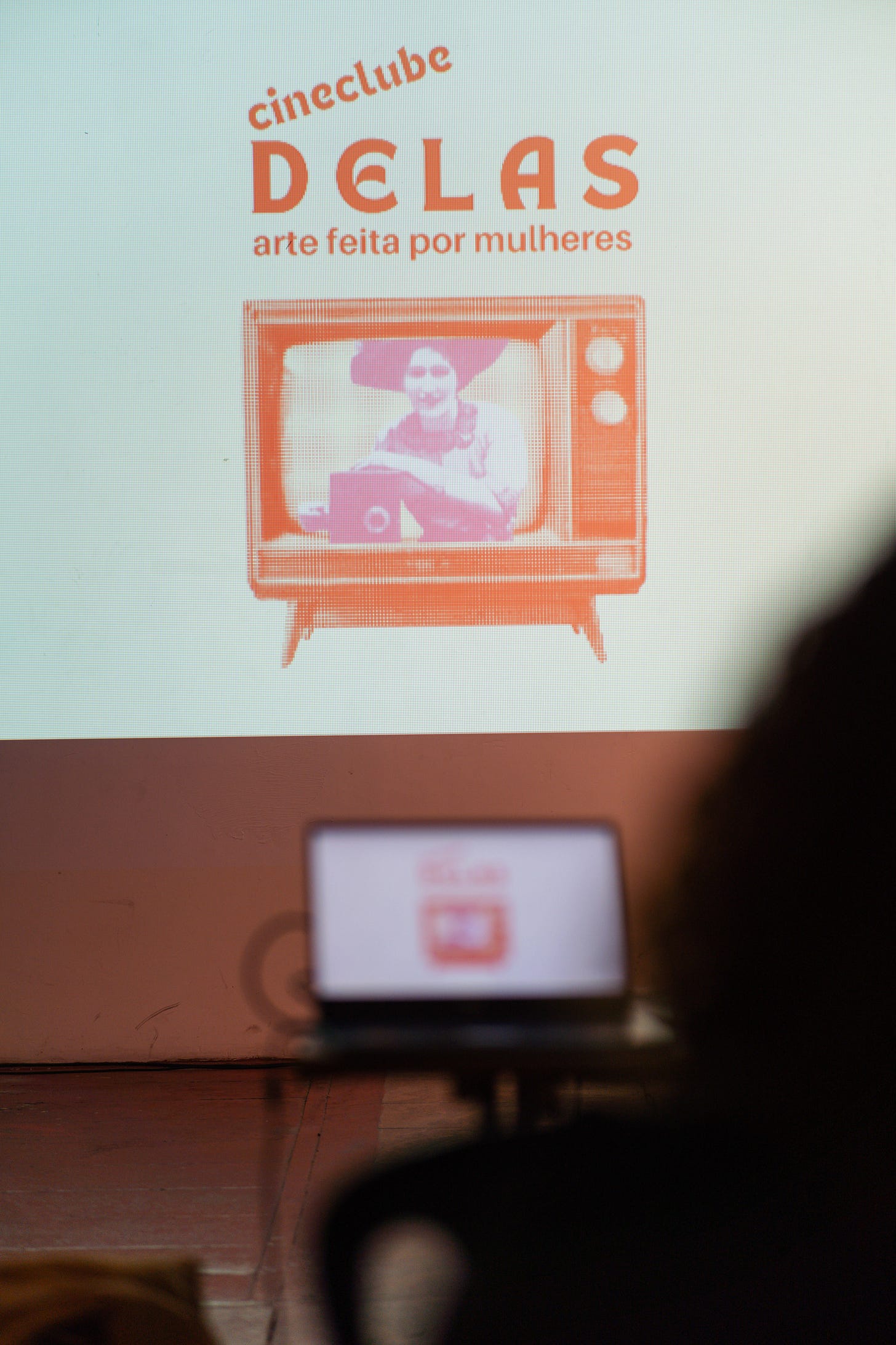 Fotografia de uma tela em branco com projeção da marca do Cineclube Delas – arte feita por mulheres, contendo o letreiro “Cineclube Delas – arte feita por mulheres” e uma televisão antiga reproduzindo na tela a imagem de uma mulher manuseando uma câmera. Mais abaixo, em desfoque, um notebook e a silhueta de uma pessoa assistindo à projeção.