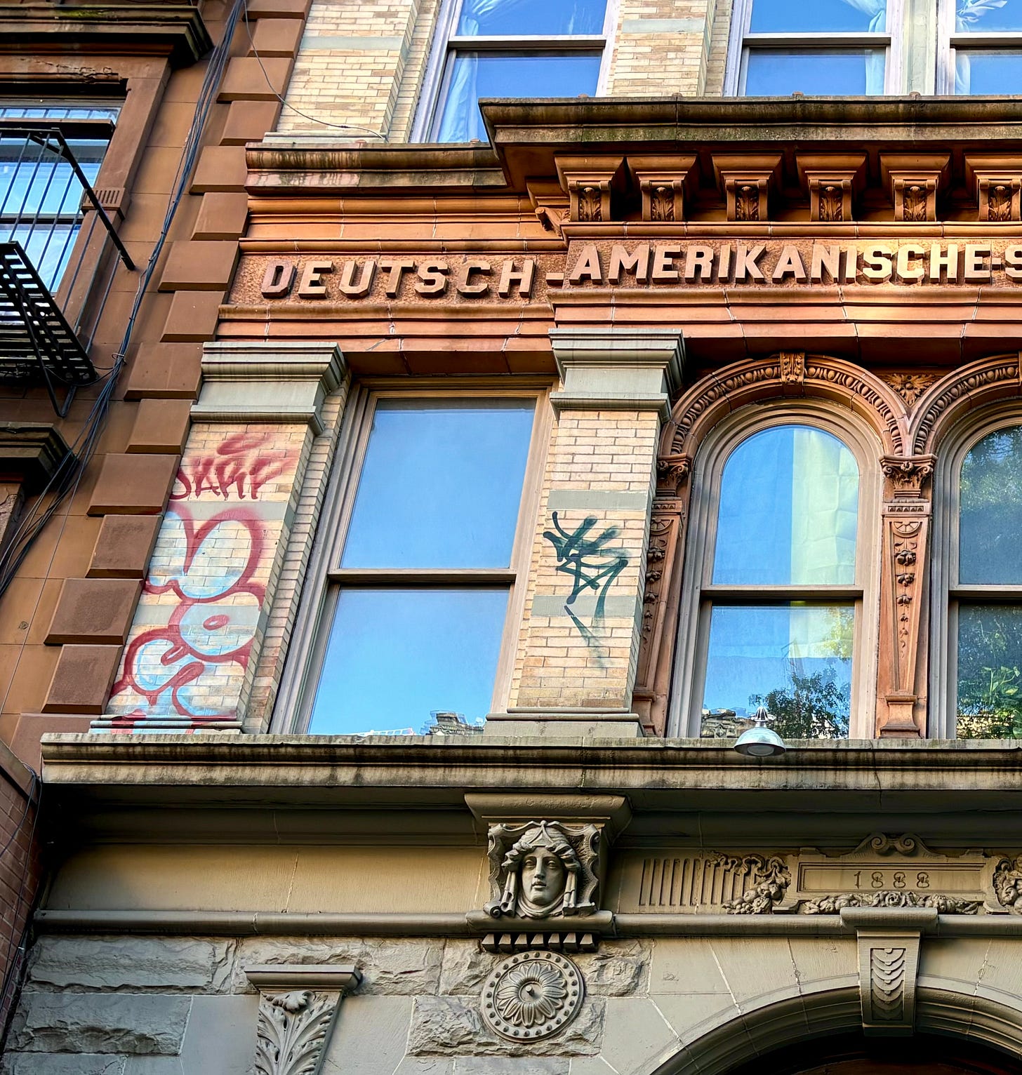 A close-up of a stone relief on a graffitied building that reads DEUTSCHE AMERIK ANISCHE-S. Below it has been marked 1888, to commemorate the year built.