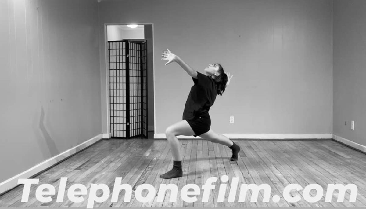 In a black and white photo, a dancer is mid-performance in a brightly-lit room with wood floor panels. Telephonefilm.com.