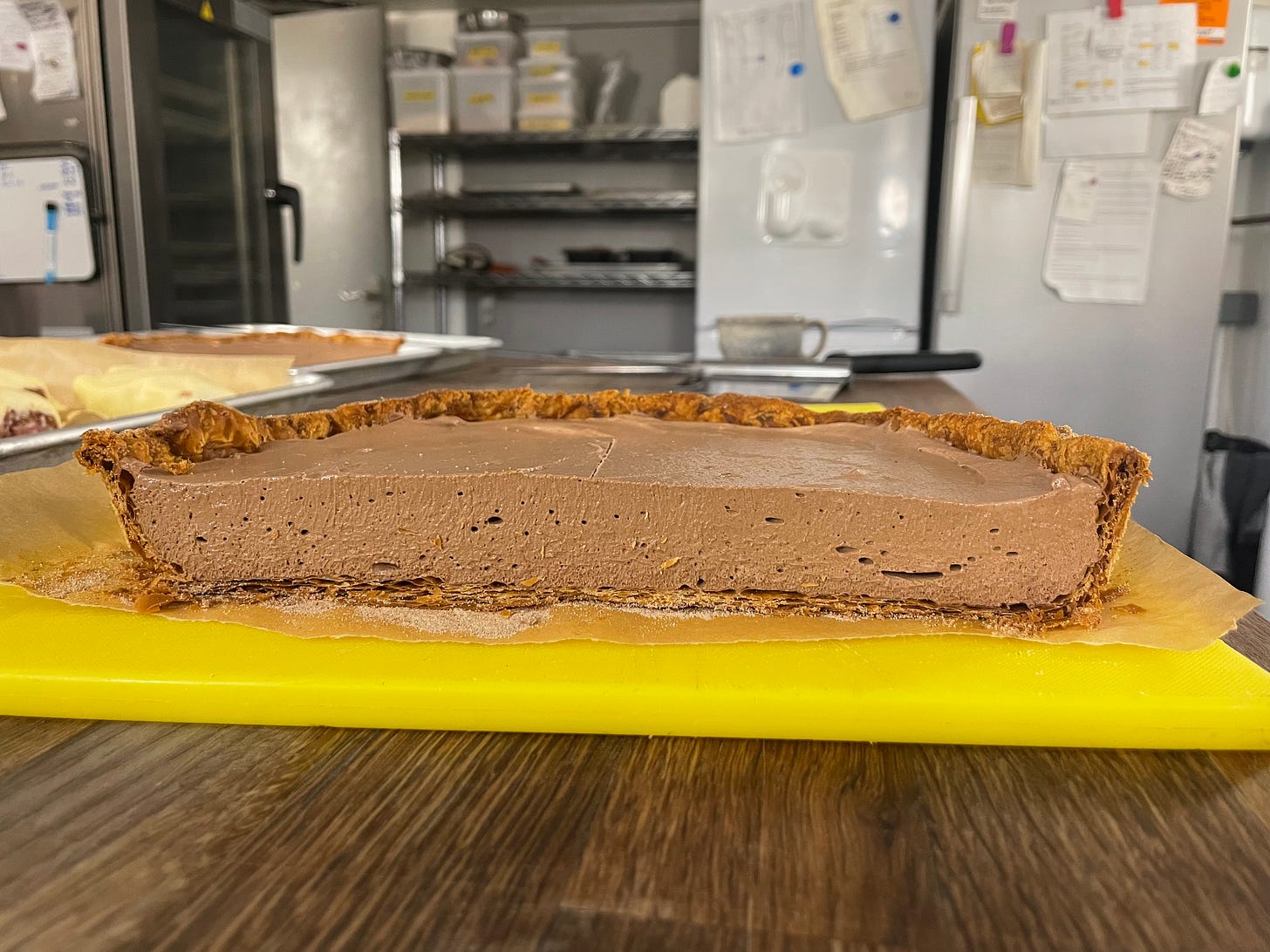 A cross-section of the champurrado pie, showing off the thick layer of set chocolate custard and the crisp brown crust. The pie is on a yellow chopping board in a professional kitchen
