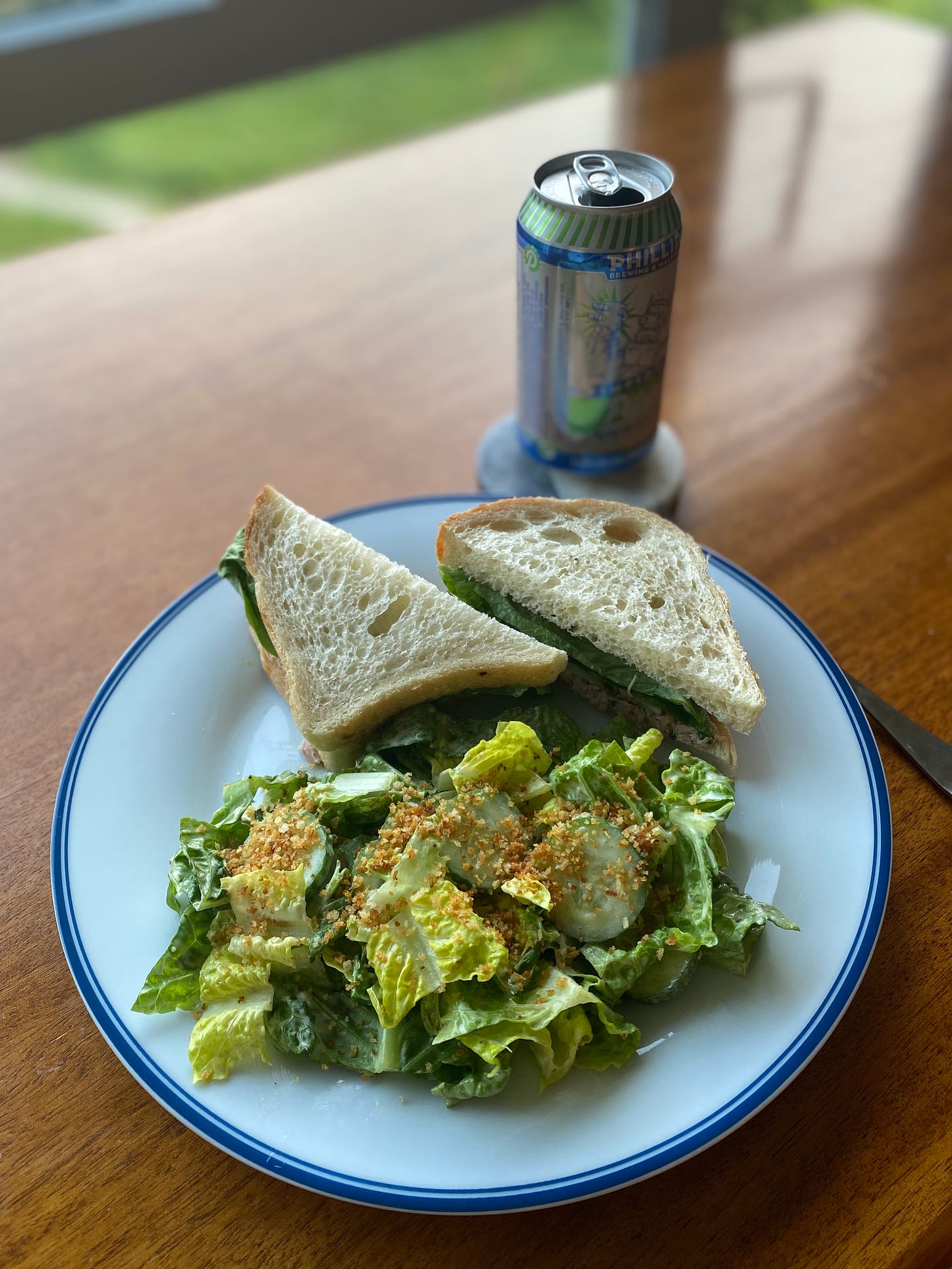 A white plate with a blue rim, filled mostly with a caesar salad of chopped romaine, cucumber, and toasted panko crumbs. At the back of the plate are two offset halves of a tuna sandwich on sourdough. In the background, a can of Phillips pilsner sits on a coaster.