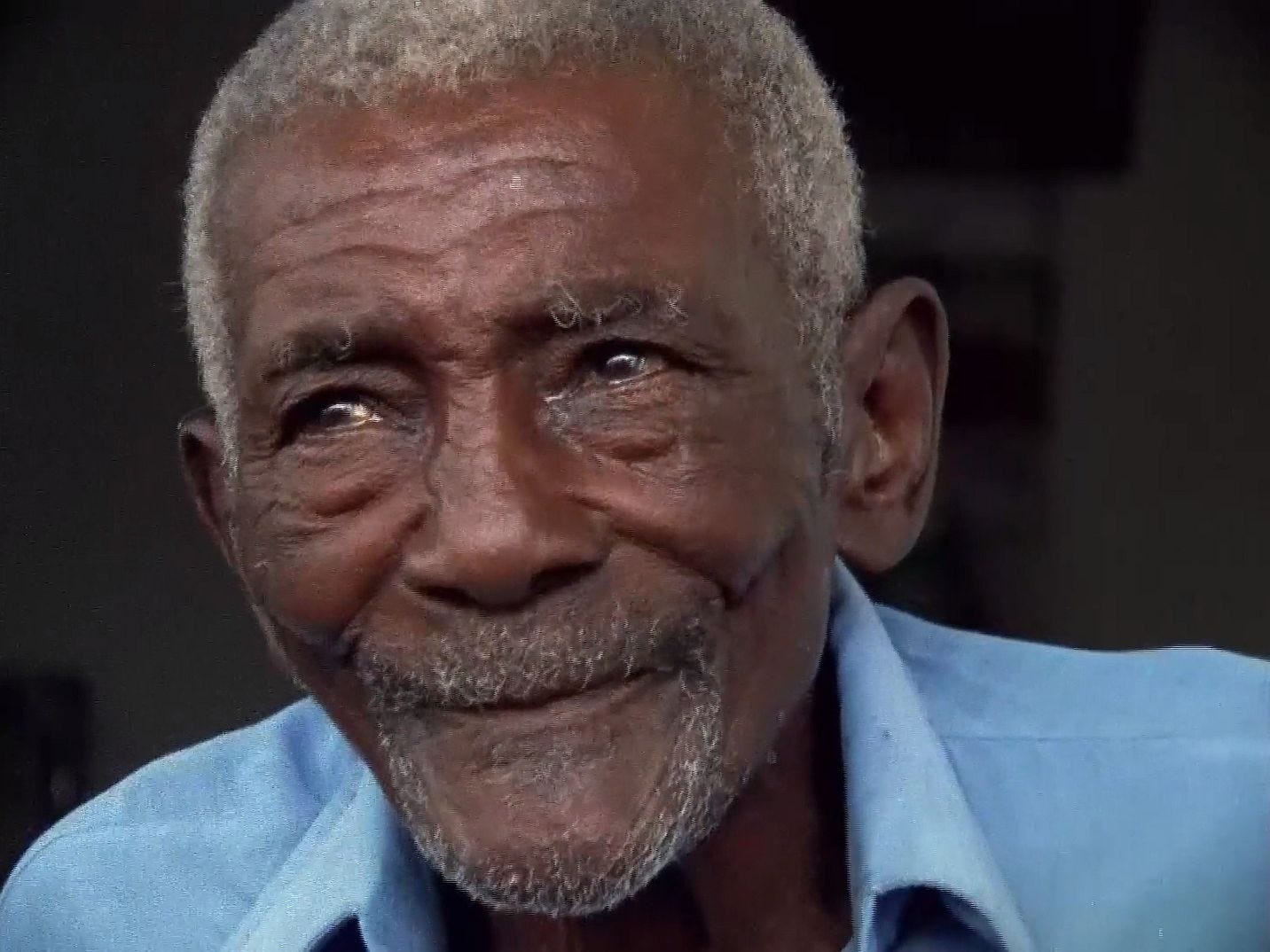 Retrato em close de um homem idoso e negro. Ele tem a barba rala e grisalha e esboça um sorriso. Usa camisa azul clara e seus olhos estão lacrimejando.
