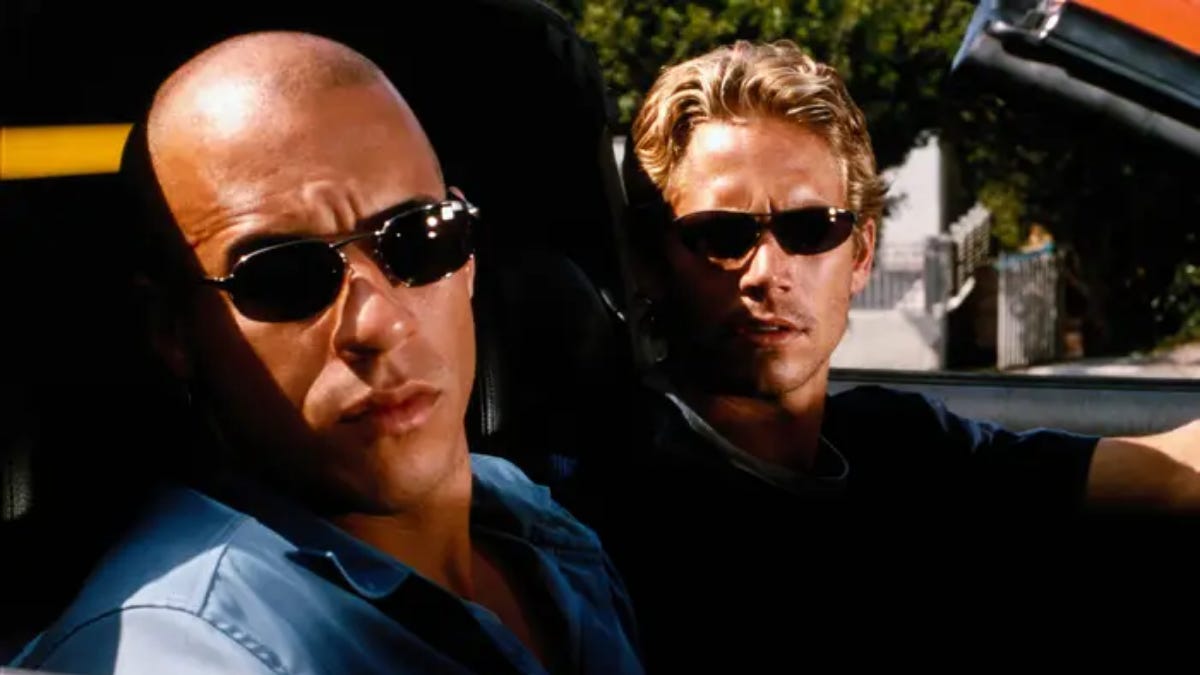Movie still from The Fast and the Furious. Two men in sunglasses sit in a convertible looking serious.