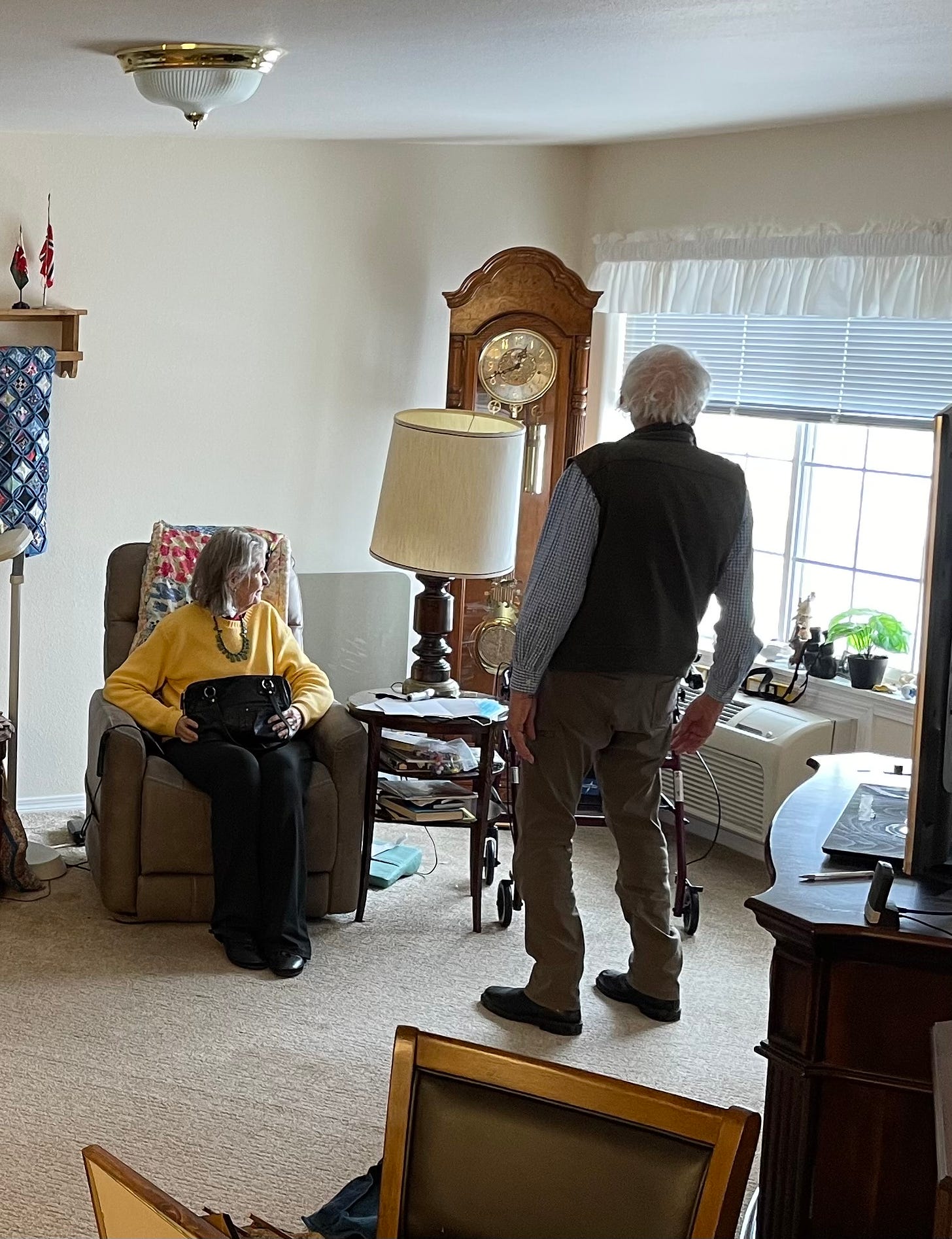 An older woman and sits and older man stands in a living room, looking out the window