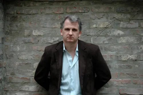 Man with gray hair wearing a white shirt and brown blazer stands in front of a brick wall.