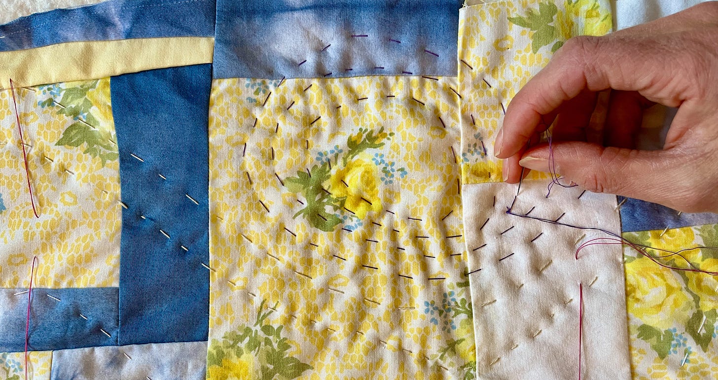 close up of my hand hand quilting a spiral on a patchwork quilt of vintage floral yellow fabric, grey fabric, and indigo tie dye. The quilt is called mediumship with my great grandmother.