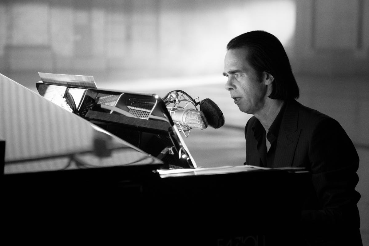 Man with shoulder length black hair sits at a grand piano, singing into a microphone.