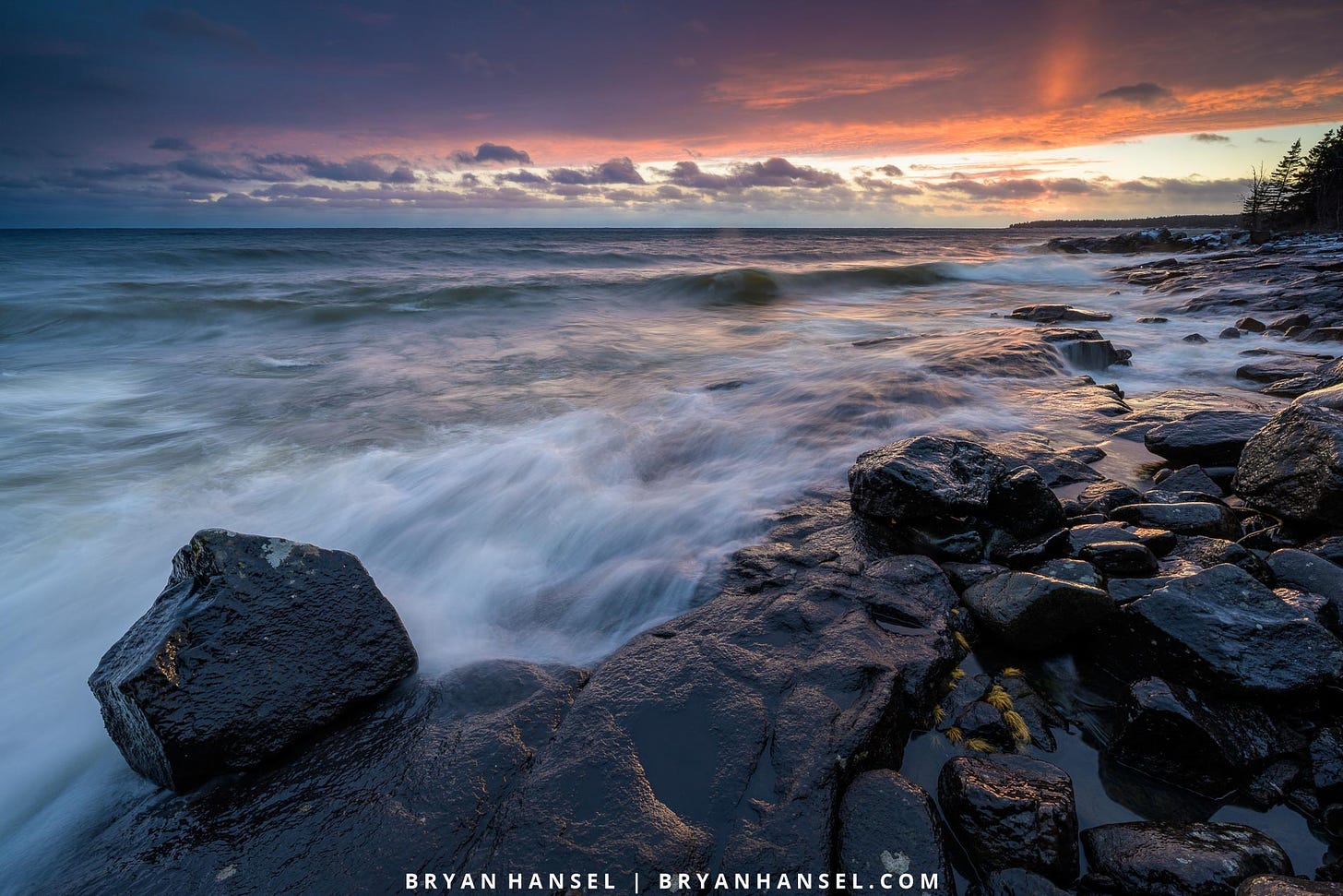 A photo of sunset over a basalt shoreline, waves and a small boulder.