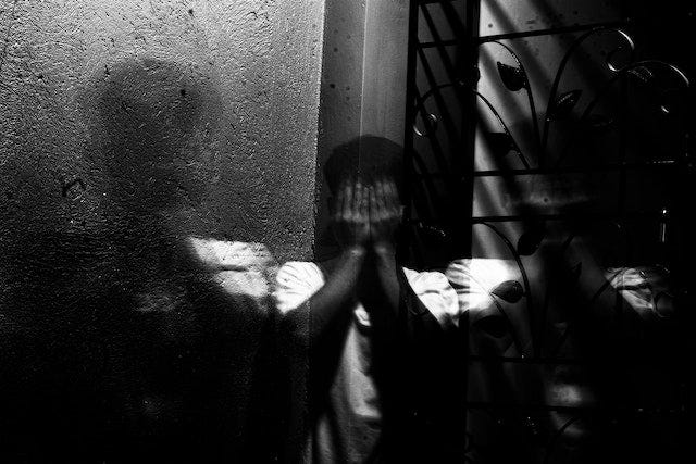 A black & white photo of a person stood in a corner, hands over their face