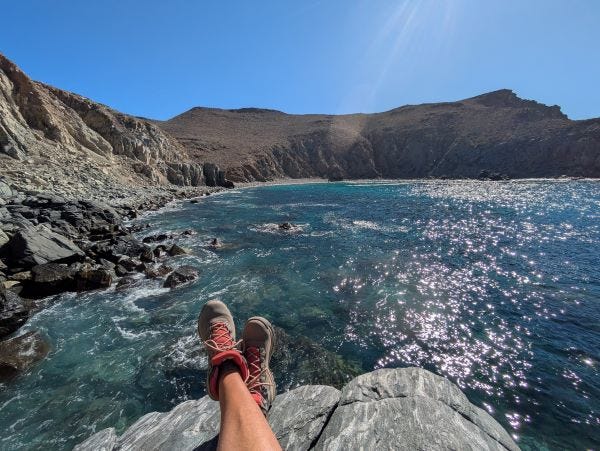 my legs, bare with hiking boots on my feet, emerging into a photo of a rugged coastline with several colors of blue and green water crashing on the rocks