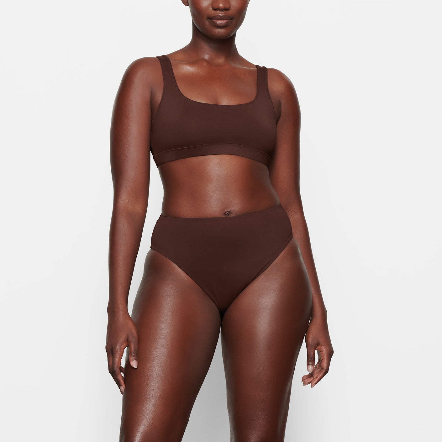 A MODEL STANDS FACING FORWARD WEARING THE SKIMS SWIM MID WAIST BOTTOM IN COCOA | SEE: M, L, XL