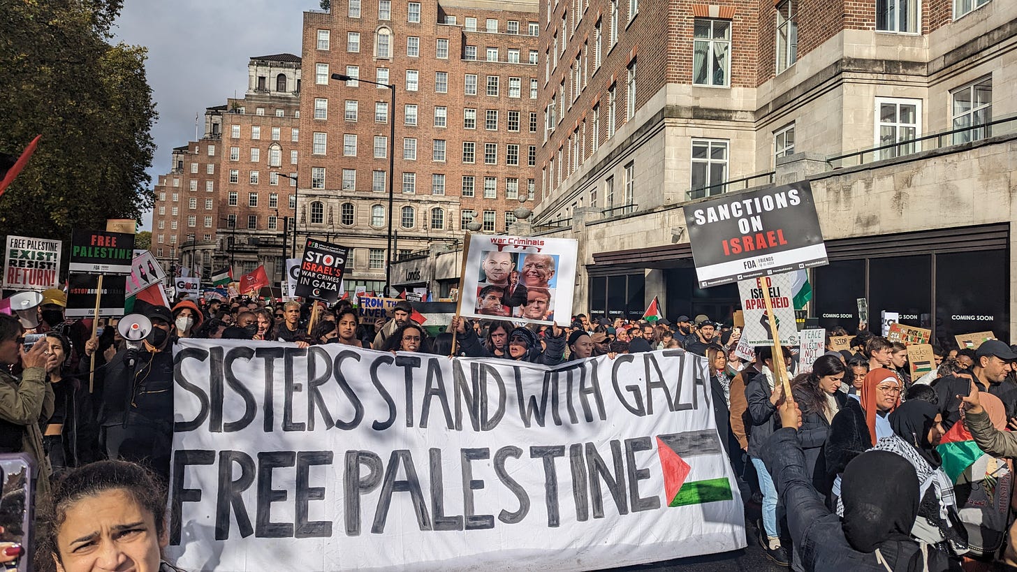 Image shows marchers at a Palestine solidarity protest in central London on 21 October 2023. At the centre of the image, people are carrying a large banner. It reads 'Sisters stand with Gaza. Free Palestine'.