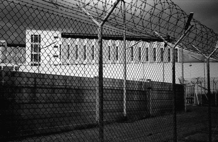 Schwarzweiß-Foto der Justizvollzugsanstalt Stuttgart-Stammheim, fotografiert von außen direkt vorm Stacheldraht-Zaun. Zu sehen sind die Mauer und ein Gefängnistrakt.