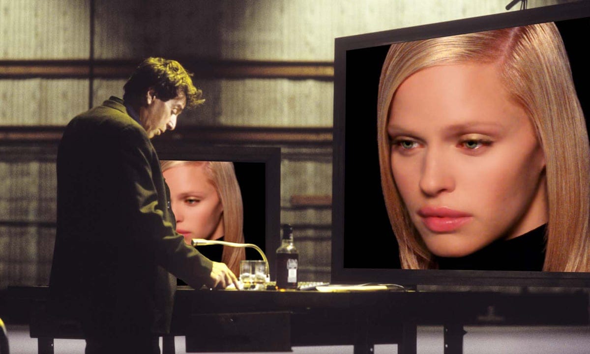 A white middle-aged man stands in front of two computer monitors sowhing the disembodied head of a white blond woman who looks digitized