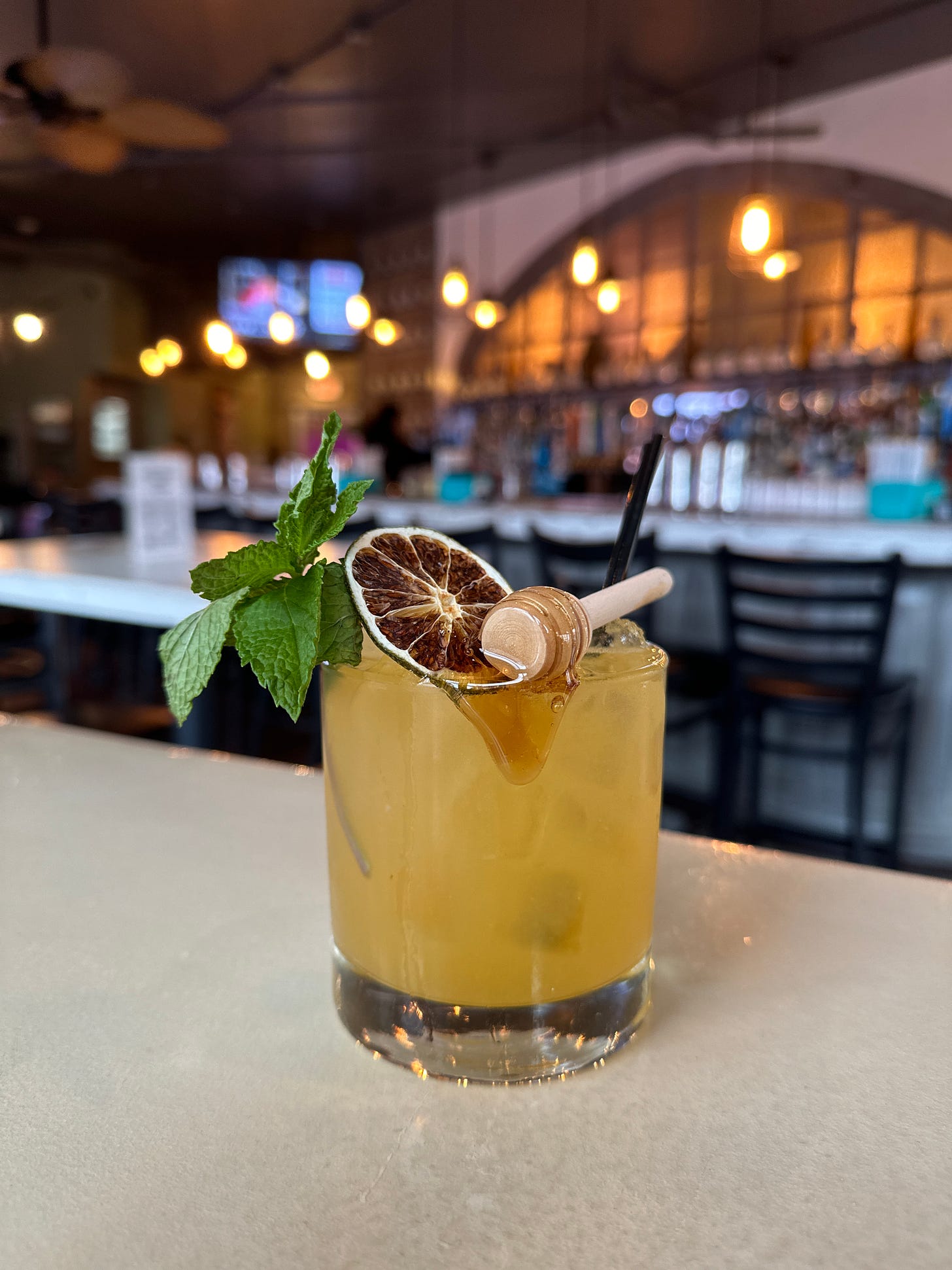 A honey whisky drink that is set against the backdrop of a restaurant bar. The drink is yellow in a whiskey glass, embellished with mint leaves, a slice of dried lime, and mini honey dipper. Honey can be seen flowing down the front of the glass.
