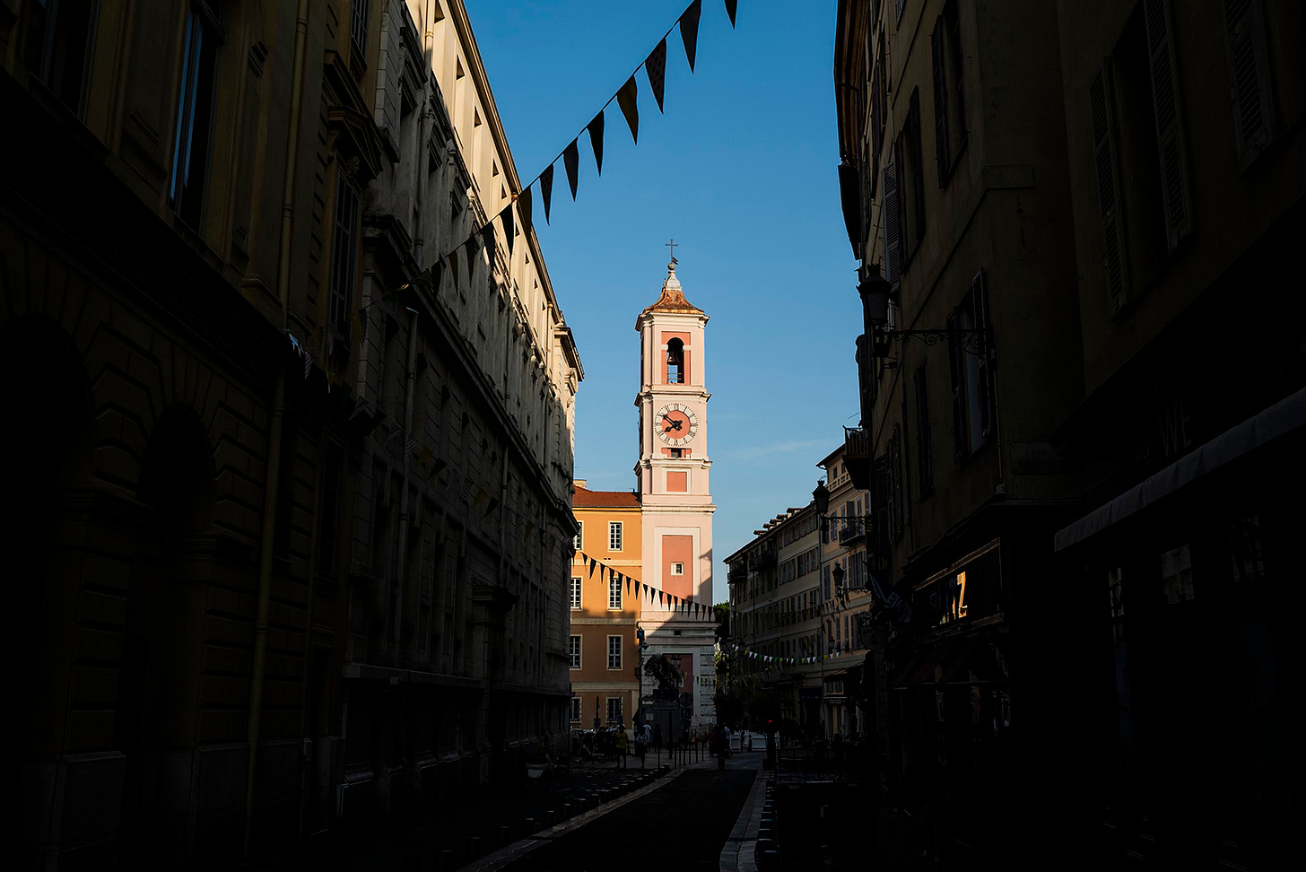 Church Tower at Sunrise in Nice – A narrow European street in early morning light, leading toward a church tower bathed in golden sunlight, contrasting with the darkened buildings on either side.