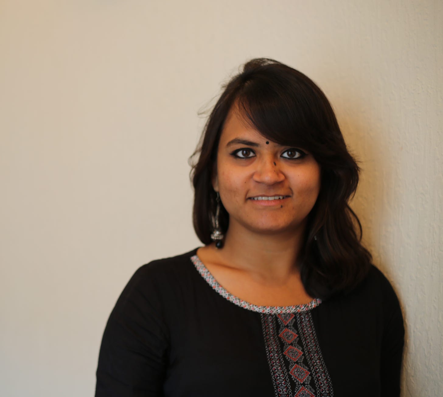 Shweta, a young Indian woman with black wavy hair falling to her shoulders is dressed in a black kurta with red and white embroidery. Shweta, who has a mole on her nose and below her lower lip is also wearing black and oxidised earrings, a black bindi and smiling into the camera.