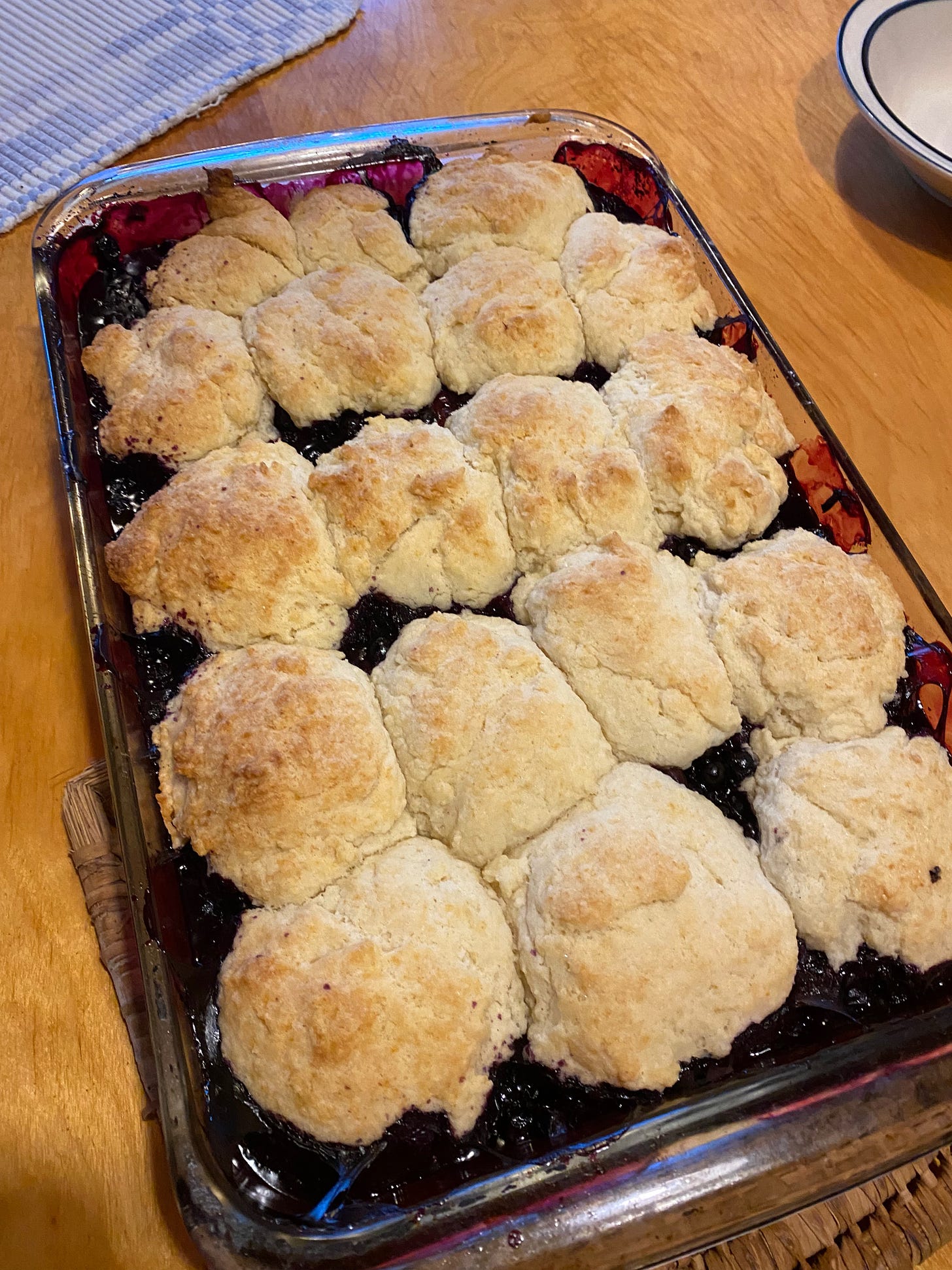 A glass baking dish of blueberry cobbler: small round mounds of golden brown biscuit topping sit atop bubbling, deep purple blueberries.