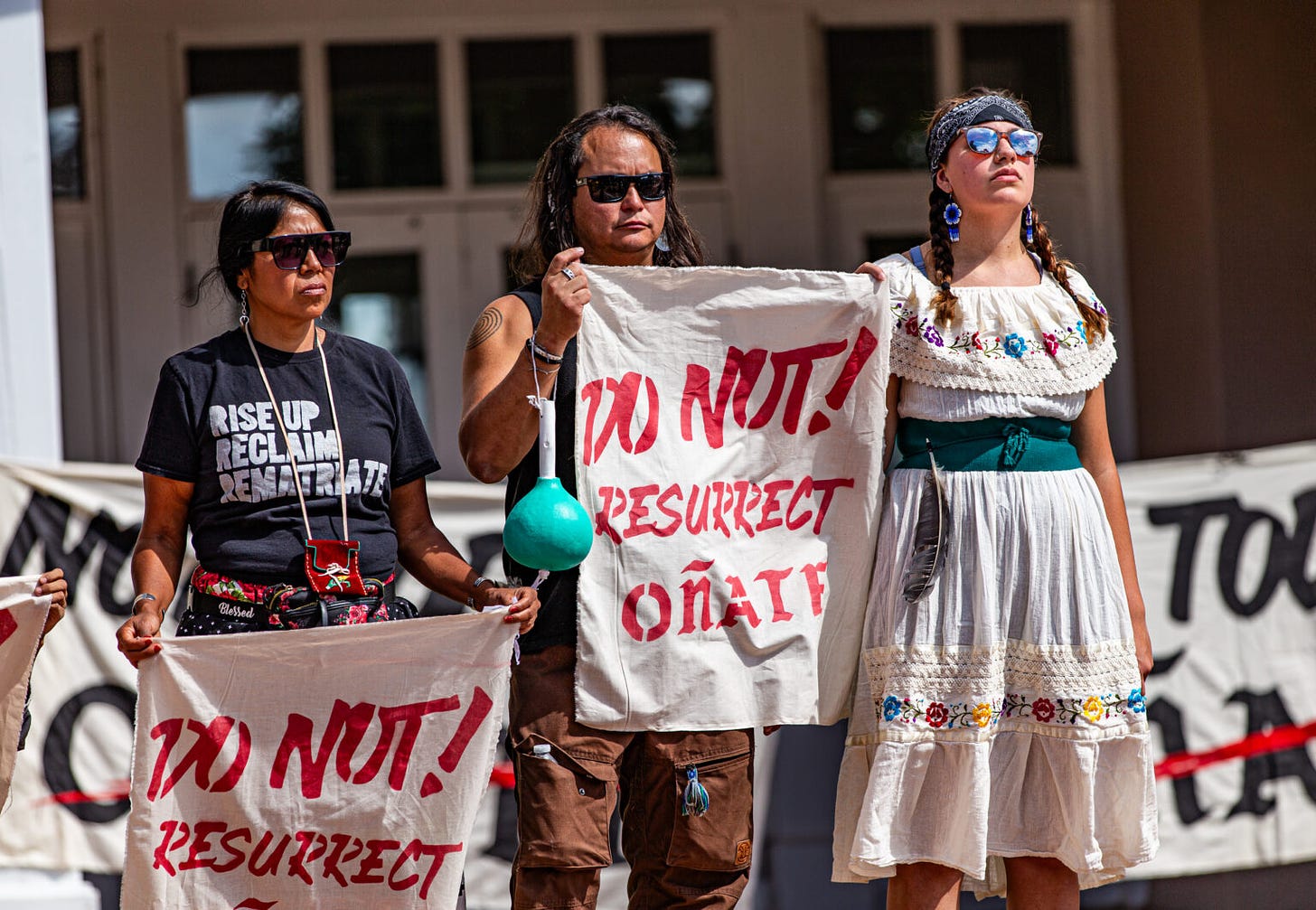 Three people stand side-by-side, some holding signs that say "Do Not! Resurrect Oñate"