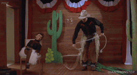A man swinging a lasso on stage beside a ventriloquism dummy on a chair.