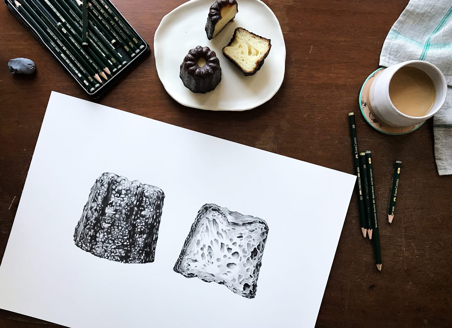 A birds eye view of a drawing of a canele pastry, with pencils to the top right, a plate with actual canele top center and a cup of coffee to the top right. Pencils and an eraser lay scattered on the wooden desk next to the drawing