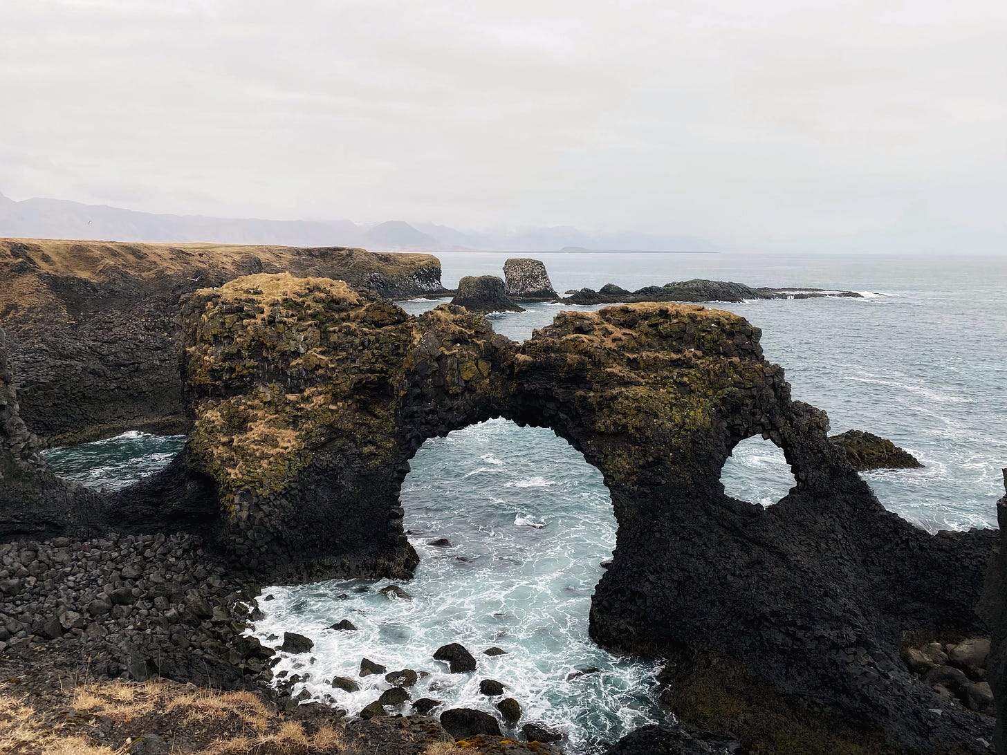 Gatklettur, a natural stone bridge over the sea.  The dark lava rocks stand out against the icy blue ocean and foamy white waves.  The top of the arch is covered with golden moss and the sky is a light gray.  Distant mountains are shown in the background.