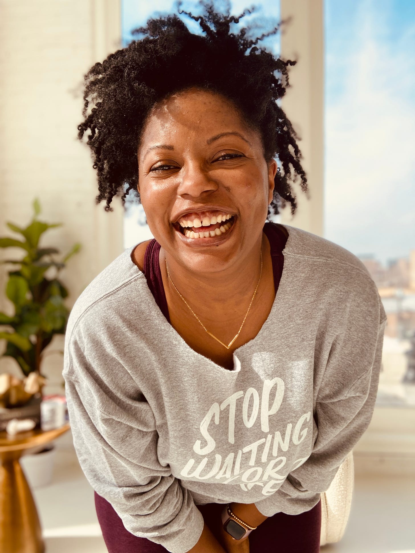 Picture of a Black woman with natural hair smiling, wearing a gold necklace, purple sports bra, matching leggings and a grey sweatshirt with white lettering that reads Stop Waiting for Perfect
