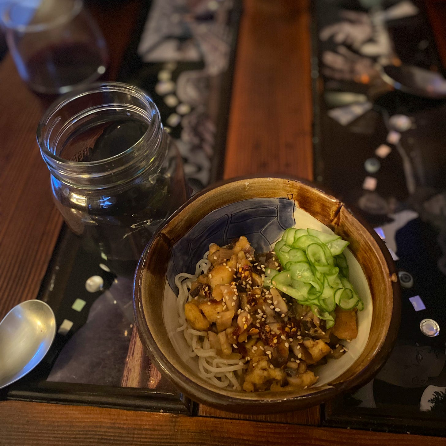 A small bowl of ramen noodles with fried tofu and mushrooms on top, covered in sauce, chili oil, and sesame seeds. A small pile of thinly sliced quick pickles rests at the edge of the bowl. Beside it sits a mason jar of dark beer.