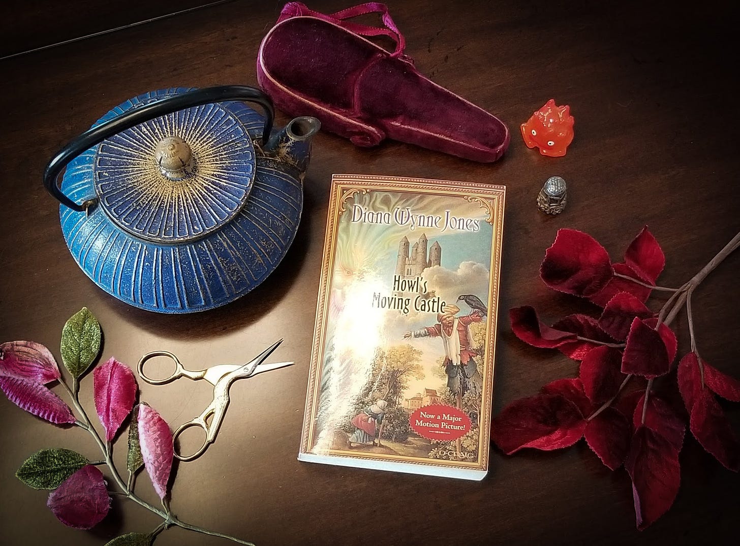 Book of Howl's Moving Castle by Diana Wynne Jones next to a blue teapot, velvet leaves, and small crane scissors.