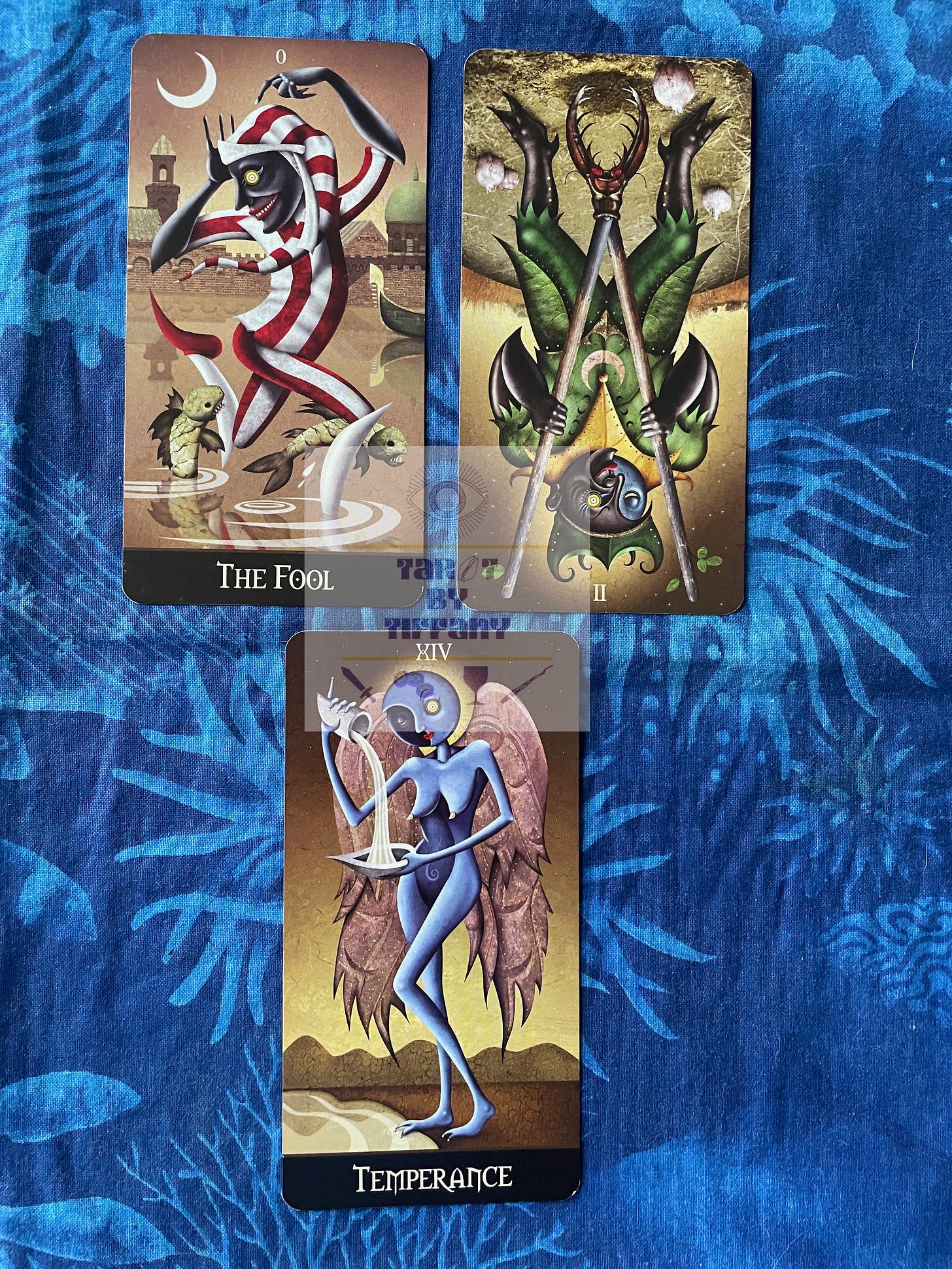 3-card reading with the Deviant Moon Tarot. The cards are laid out on a cobalt blue cloth with an aquatic motif in lighter blue. Top cards L-R: The Fool, 2 of Wands reversed. Bottom card: Temperance.