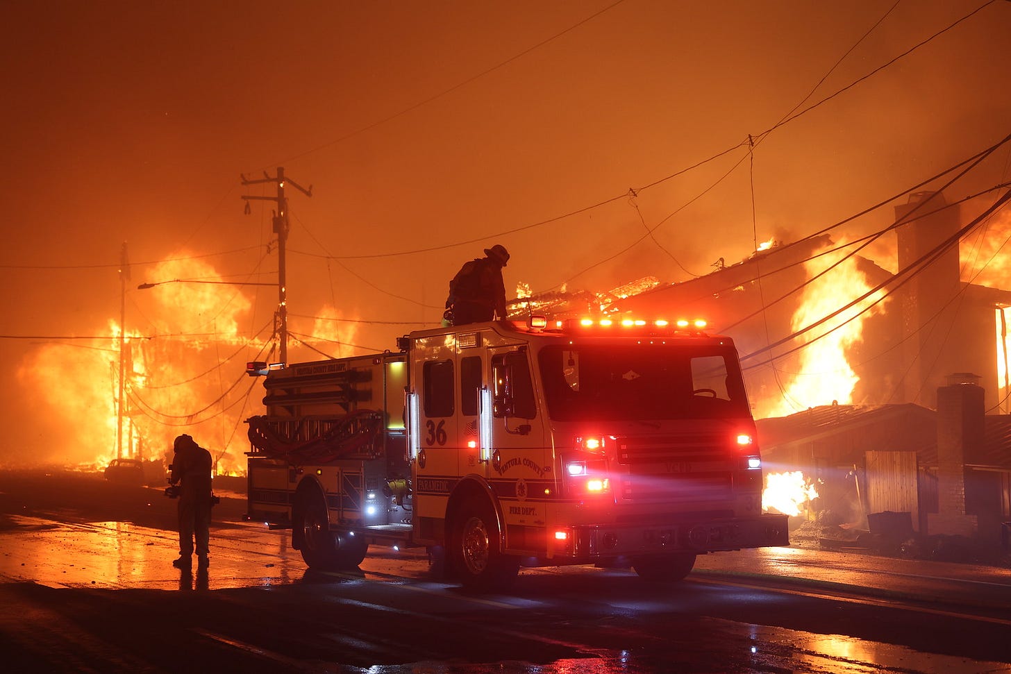 Two tired firefighters by a firetruck. Buildings and power lines in the background are on fire.