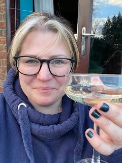 Me, facing the camera, and raising a coupe glass of prosecco. I'm smiling, wearing a blue hoody, and have green sparkly nail varnish.