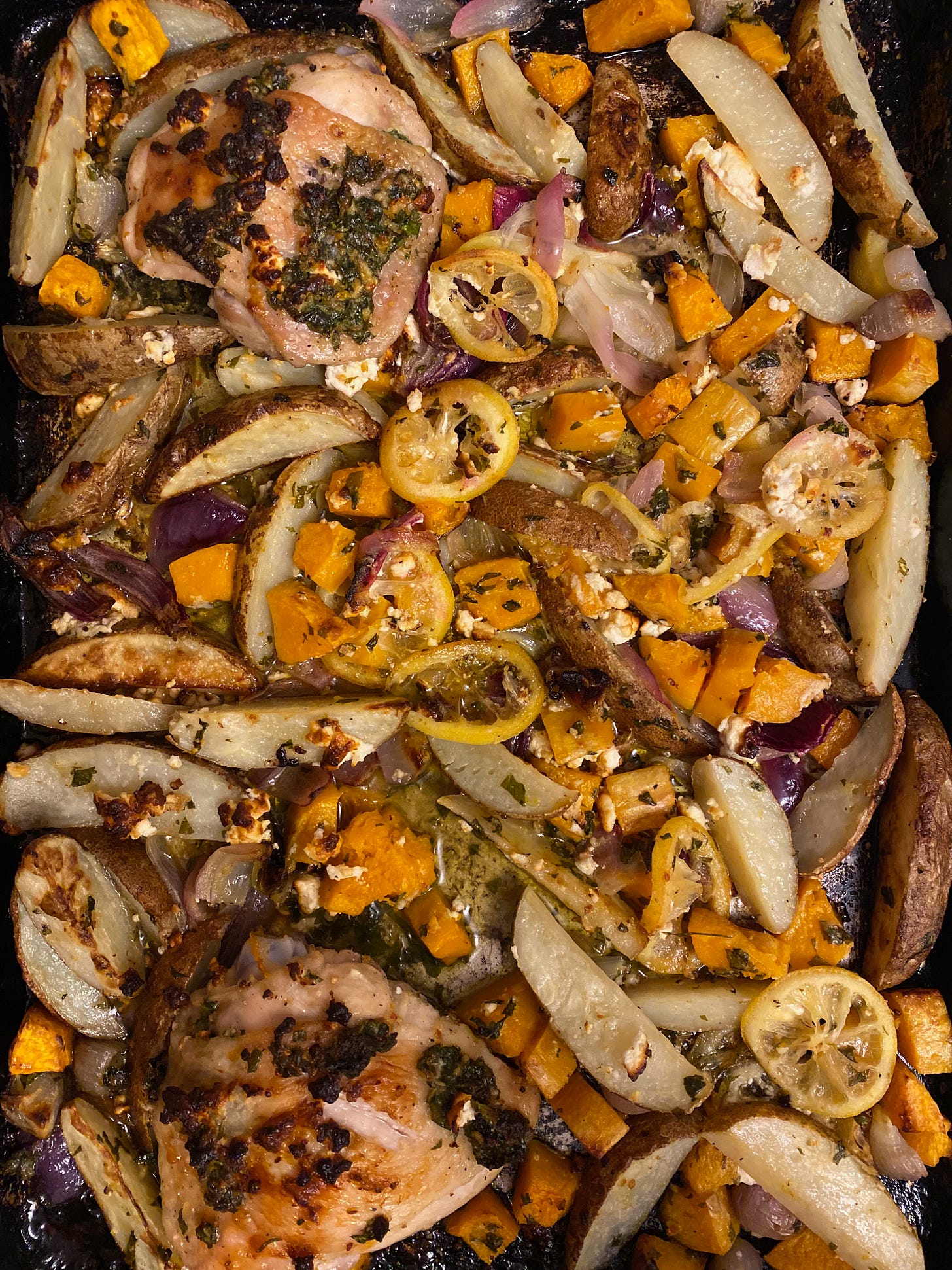 Close-up of a sheet pan full of roasted potatoes and sweet potatoes, sliced lemons, chunks of feta, and chicken thighs.