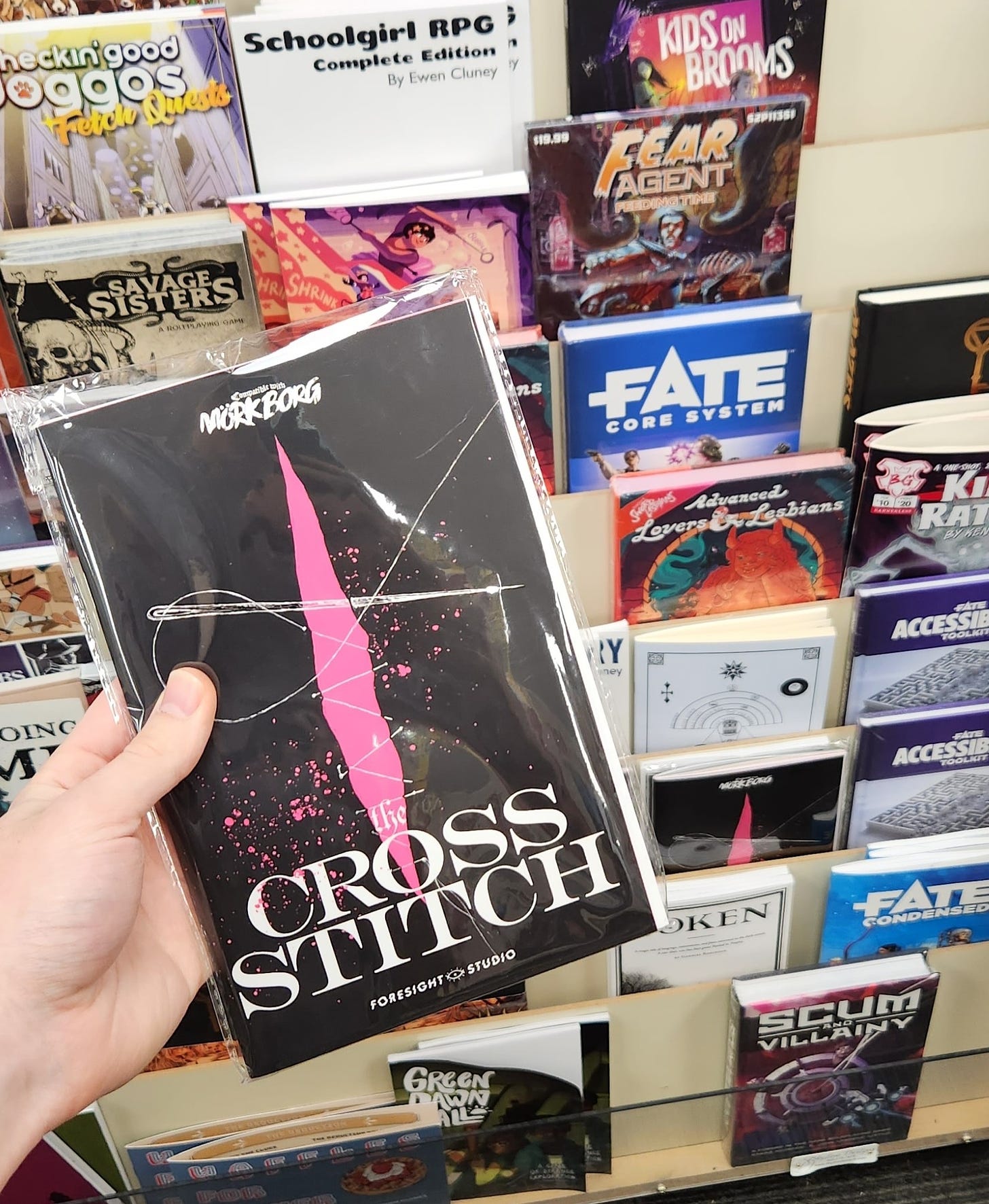 A hand holding a copy of Mork Borg zine The Cross Stitch, in front of a rack of other TTRPG books and supplements.