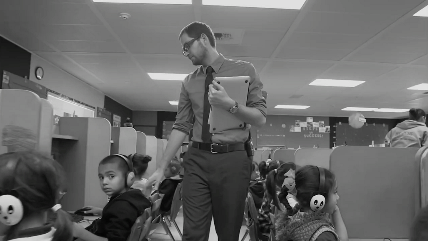 An image of a teacher walking between rows of laptops, supervising student computer use.