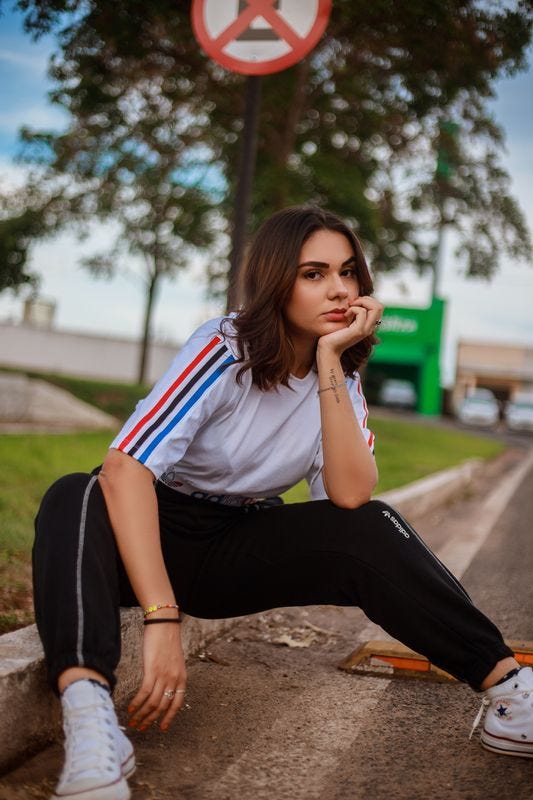 Light-skinned person wearing athletic  clothes, sitting on a sidewalk curb and scowling