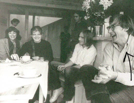 A group of people sits around a table set with teacups and saucers. Christopher Alexander sits on the right, clapping his hands and laughing as if at a hilarious joke.