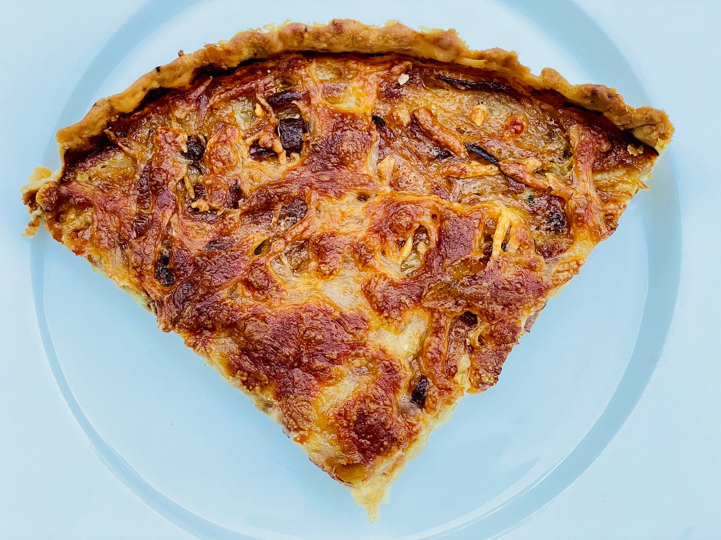 A close-up image of a single slice of Tarte à l’oignon Alsacienne, showing the golden-brown cheese and onion filling on a light, flaky crust. The tart is served on a light blue plate. A close-up image of a single slice of Tarte à l’oignon Alsacienne, showing the golden-brown cheese and onion filling on a light, flaky crust. The tart is served on a light blue plate.