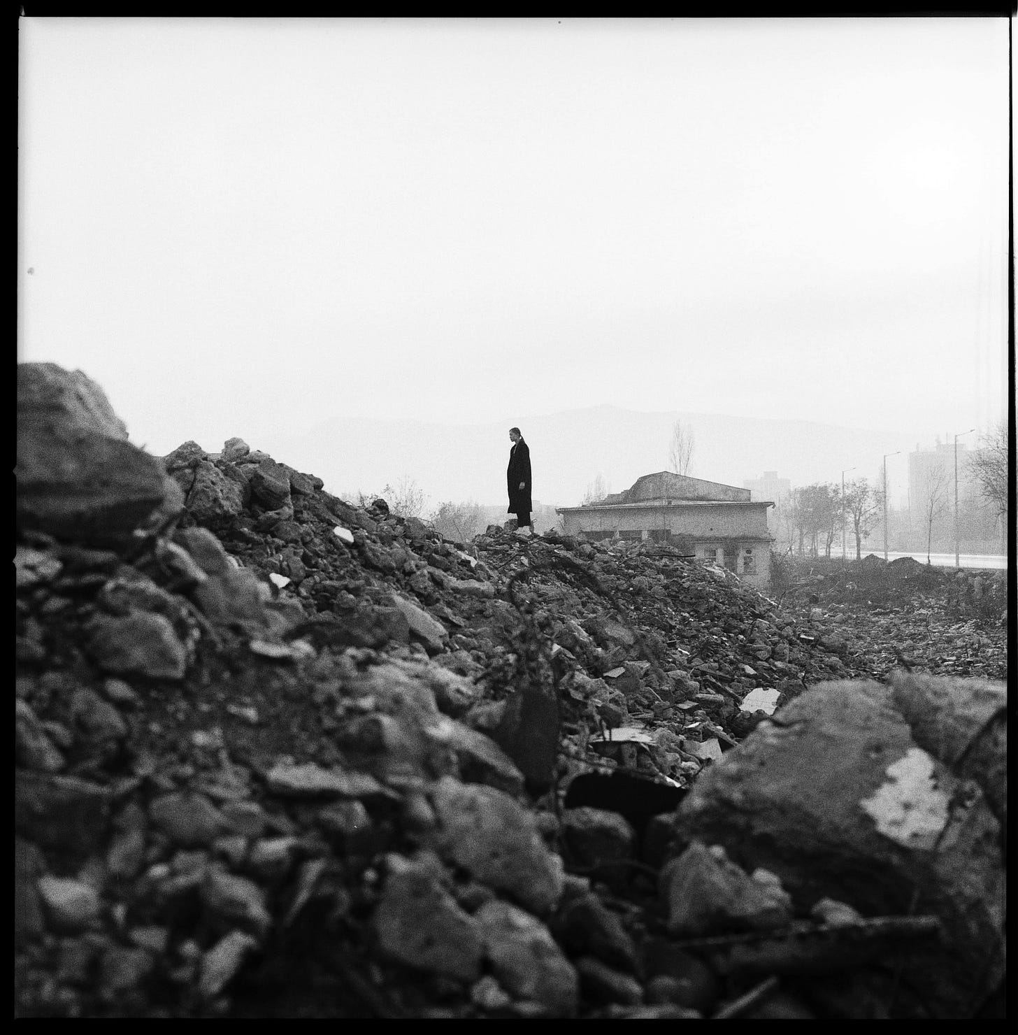 Black-and-white image showing rocky rubble in the foreground, a silhouetted and black-clad figure standing on the rubble at about the center of the image, and a partially ruined house in the background. There are trees framing the right side of the house.