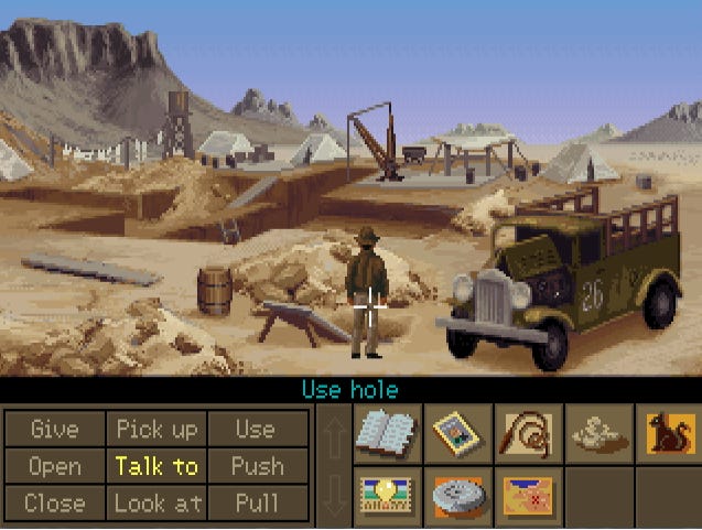 A screenshot from Indiana Jones and the Fate of Atlantis, that shows Indy out in the desert at a dig site. The verb toolbar is displayed in the bottom left, with "Use" selected, and the cursor hovers over a hole in the ground that's hidden behind Indy's body, leading to the potential command "Use Hole"
