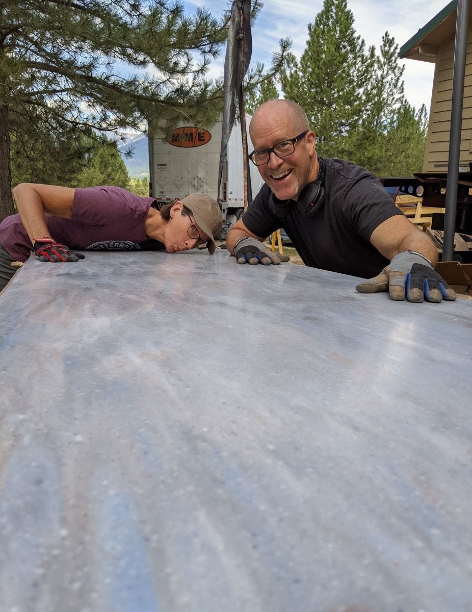 Sherry and Andy showing love for a sheet of countertop material, with Sherry leaning down to kiss it and Andy smiling with his hands caressing it