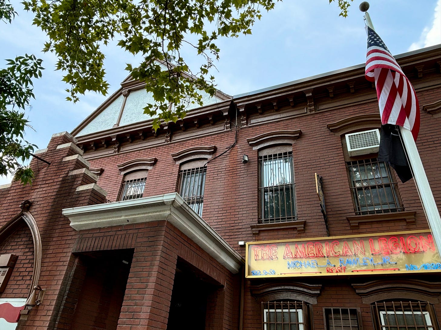 A brick building with an American flag outside. A neon sign reads The American Legion Post 1636, Michael Rawley.