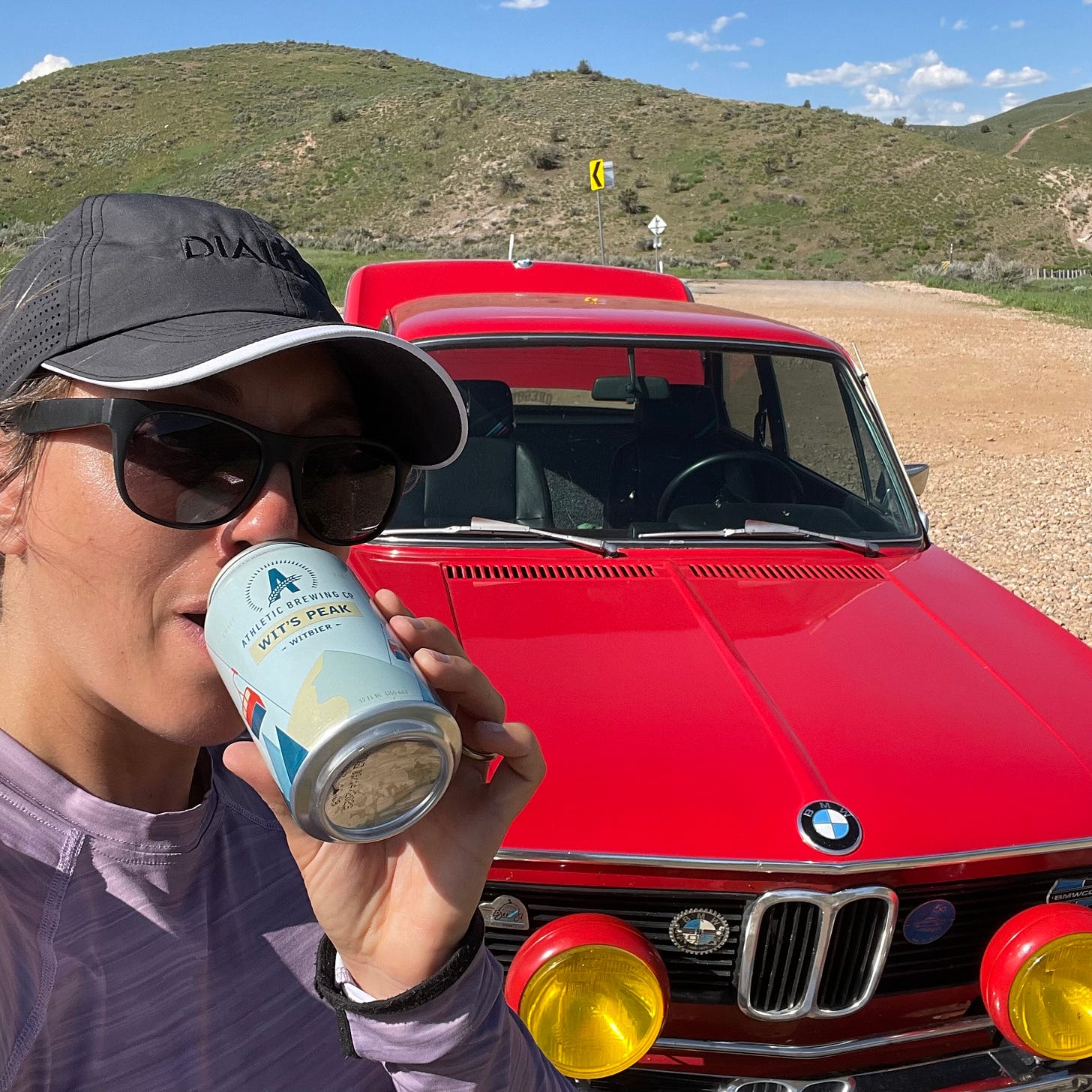 Mina drinking an Athletic Brewing Wits Peak from a can, posing next to a red antique BMW from 1976 -- the 2002 model -- with grassy hills in the background.