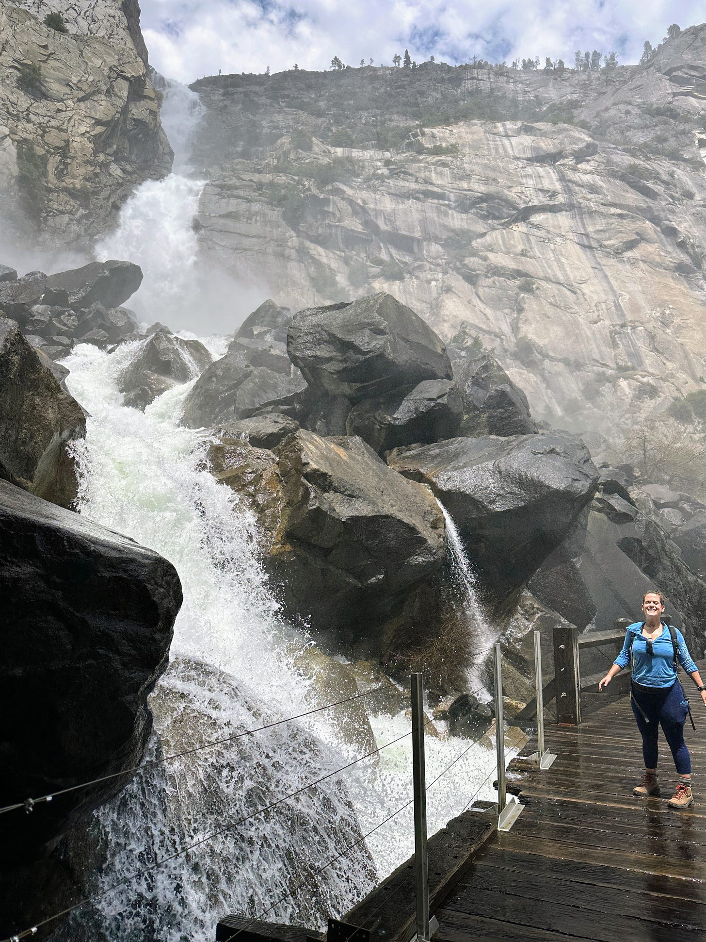 white woman in black yoga pants and blue long sleev shirt standing on a bridge by a roaring waterfall in Yosemite. She is wet and smiling
