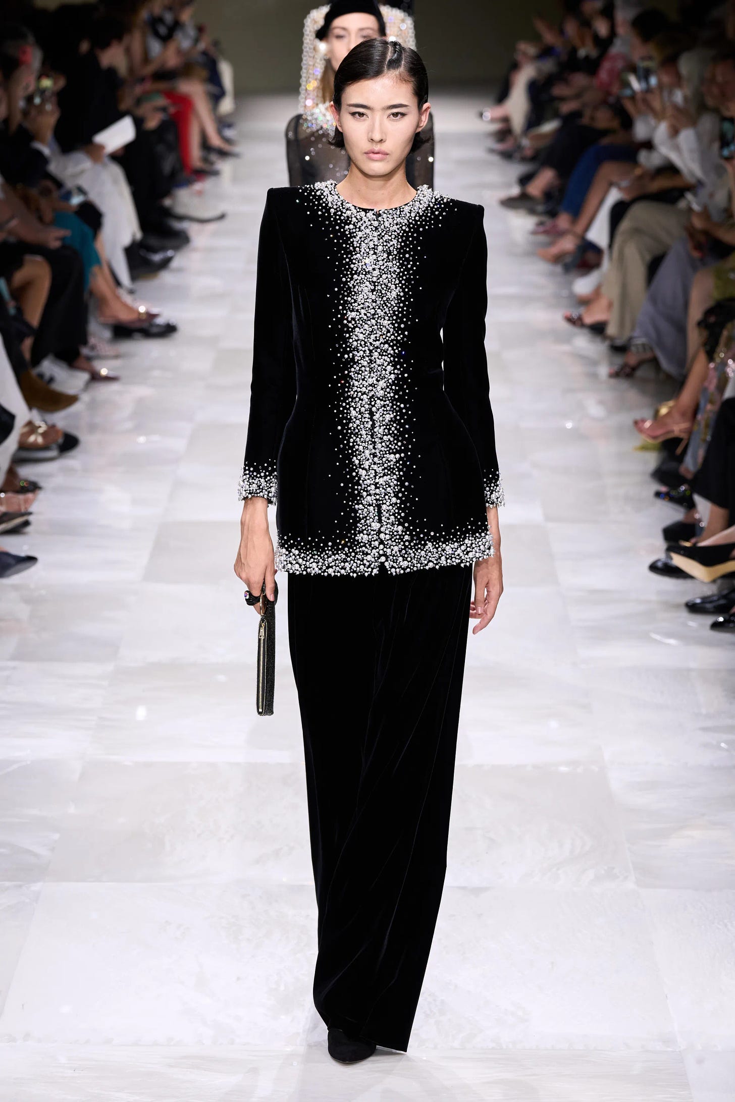 An Asian female model walks down the runway wearing a black velvet pantsuit with twinkling embroidery around the neckline, front closure, and cuffs of the jacket. 