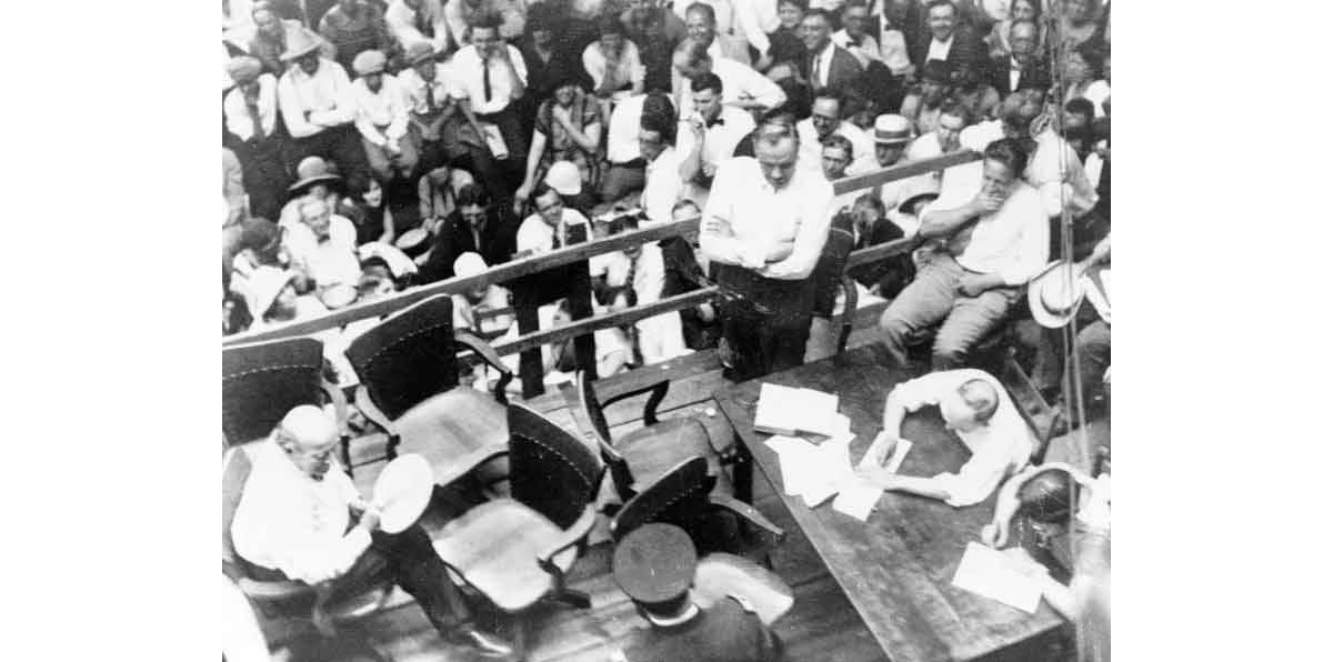 People surrounding a wooden stage with railing, William Jennings Bryan seated at left from overhead view, Clarence Darrow standing at right, with table and other court officials.