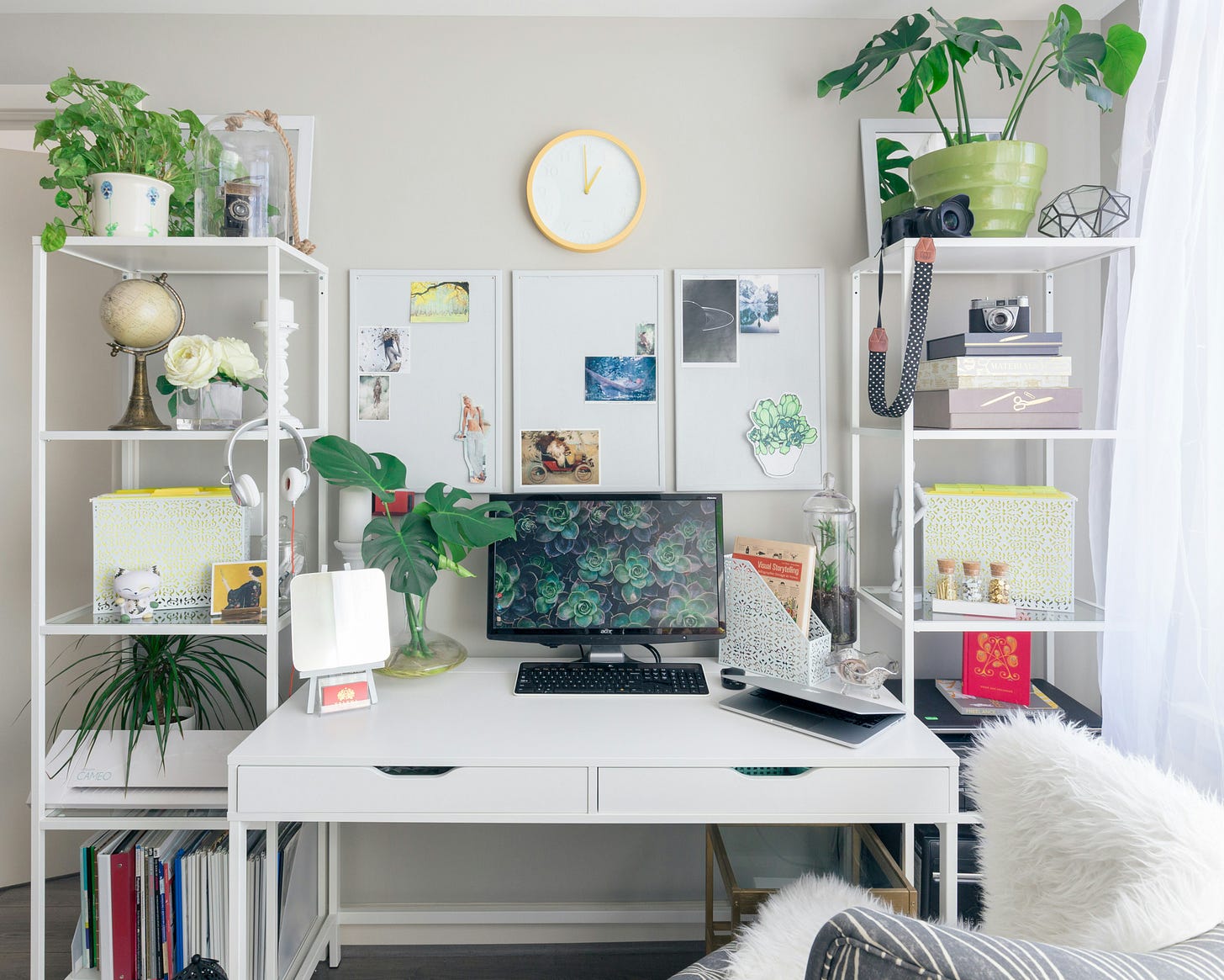 Desk with book shelves on either side in front of white boards and a clock. Desk has a computer, plants, and notebooks and bookshelves are filled with trinkets.